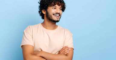 Handsome positive arabian or indian curly-haired millennial guy in a t-shirt, standing over isolated blue background with crossed arms, looking away, smiling happily